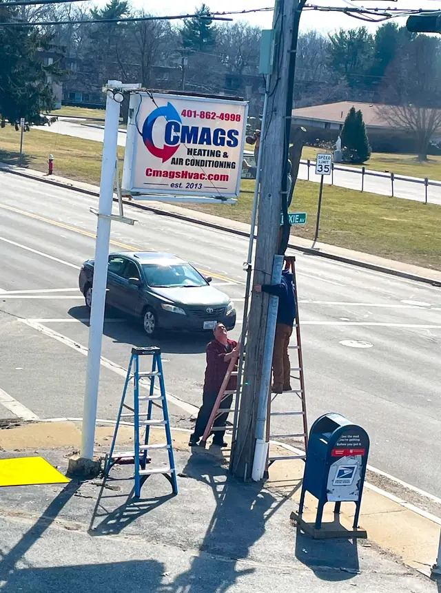 Two Cmags technicians on ladders installing the Cmags Heating and Air Conditioning roadside sign at a street corner, est. 2013 on the sign face